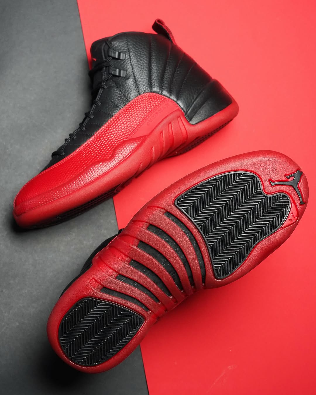 Air Jordan 12 Retro Flu Game featuring black tumbled leather upper with vibrant red textured mudguard and red outsole, displayed on a contrasting black and red background