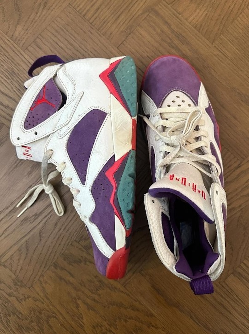 A pair of Air Jordan 7 "Joker" Sample sneakers displayed on a wooden floor, showcasing their white leather and purple suede combination.