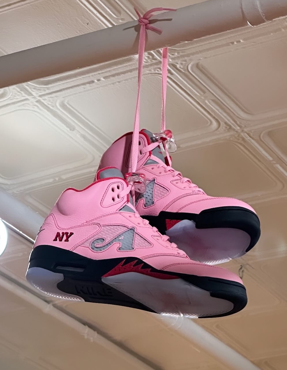 Awake NY x Air Jordan 5 collaboration featuring a vibrant pink upper, grey reflective mesh panel with an 'A' logo, 'NY' embroidery on the heel, and black/red midsole details, hanging by pink laces.