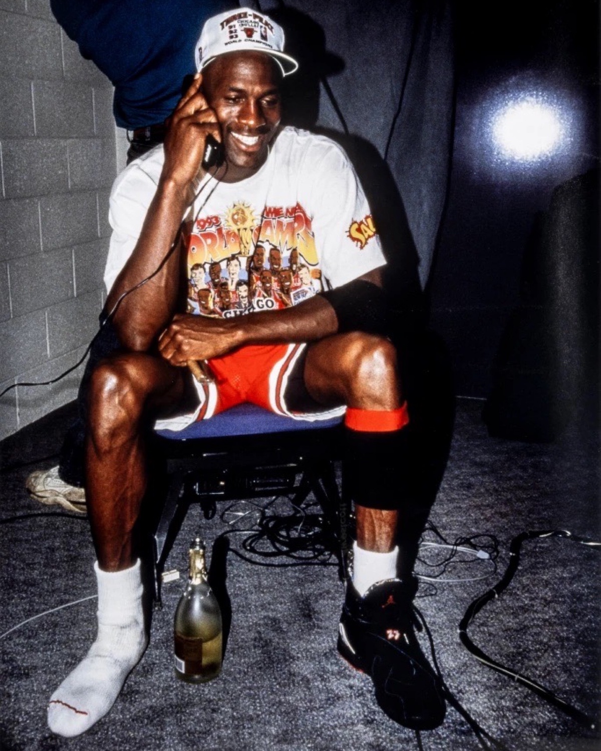 Michael Jordan celebrating the 1993 NBA Finals victory wearing a Chicago Bulls championship t-shirt, championship cap, and Air Jordan 8 sneakers. He is smiling while talking on the phone, with a champagne bottle at his feet, sitting in the locker room post-game.