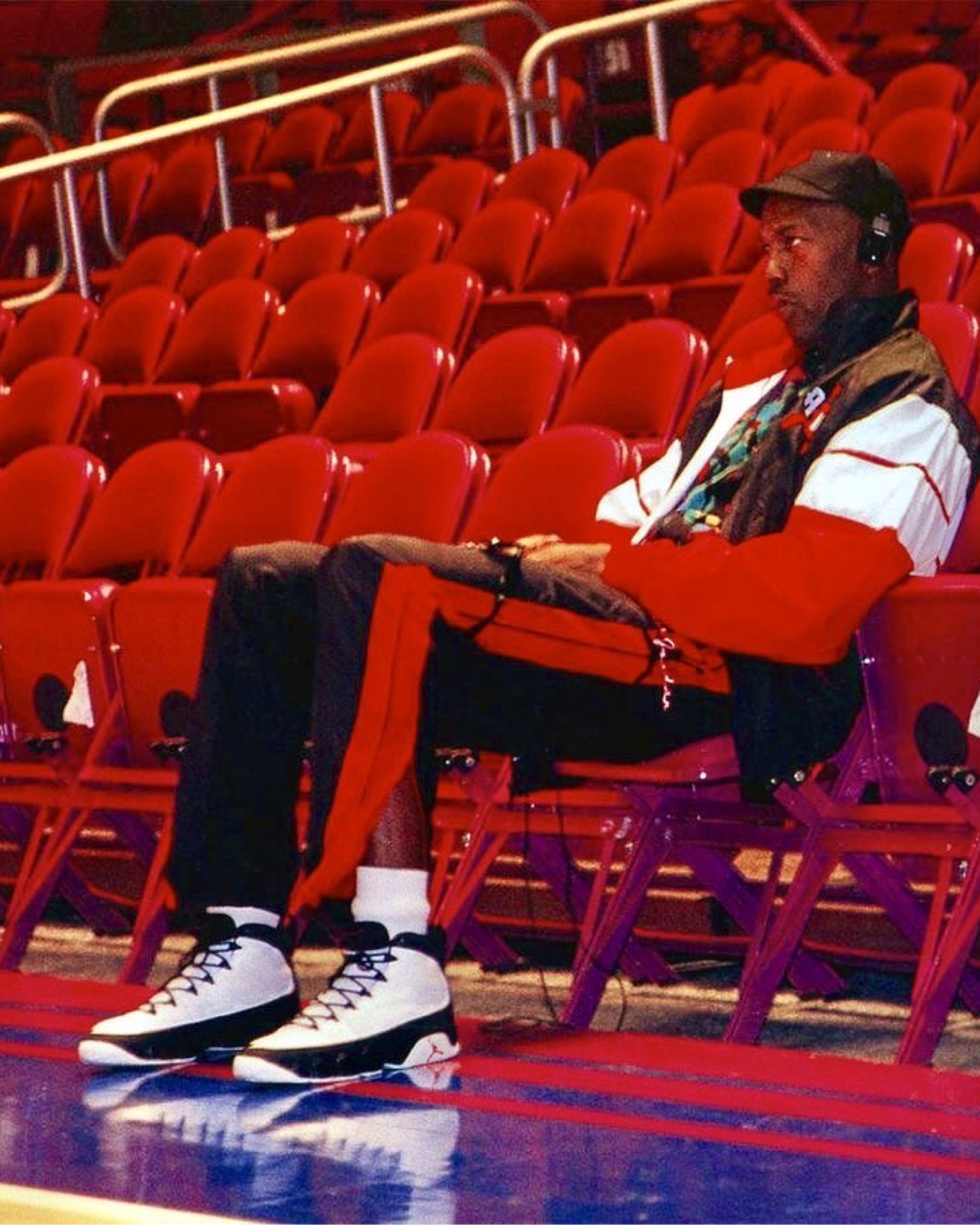 Michael Jordan sitting courtside wearing a red, black, and white tracksuit with the Air Jordan 9 'White/Black/True Red,' listening to music on headphones in an empty arena.