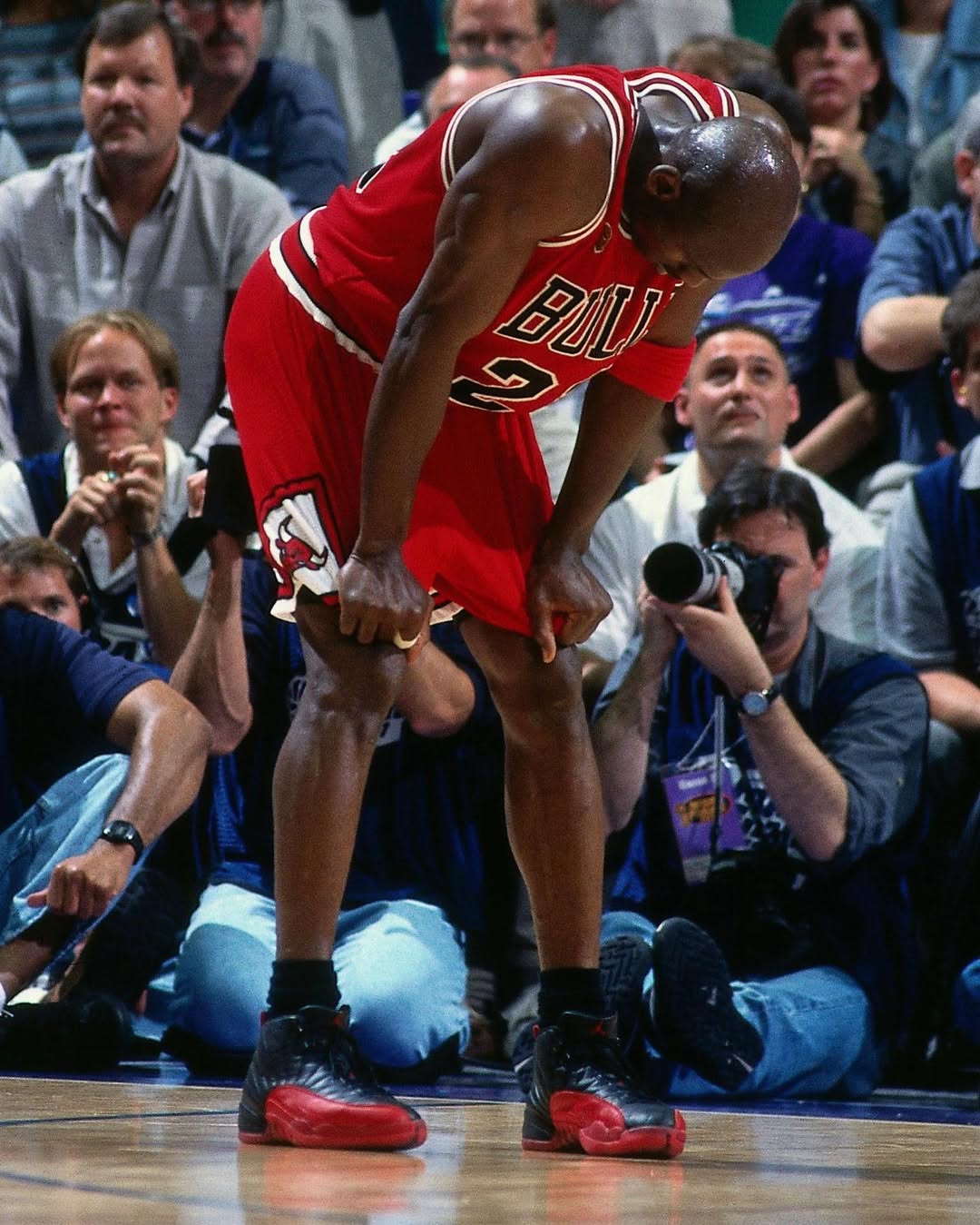Michael Jordan wearing the Chicago Bulls red jersey, bending over in exhaustion during the iconic 1997 NBA Finals Flu Game against the Utah Jazz, showcasing the black and red Air Jordan 12 Flu Game sneakers on the court with photographers and fans in the background.