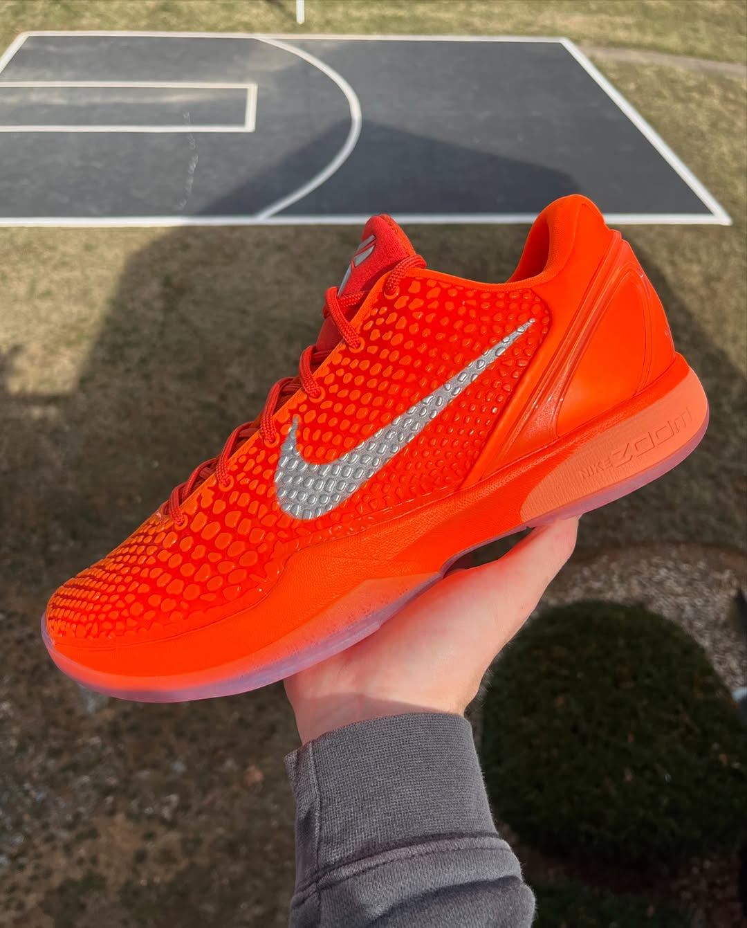 A close-up of the Nike Kobe 6 Protro "Total Orange" sneaker featuring a vibrant orange upper with a scaly texture, a metallic silver Swoosh, and a translucent sole. The shoe is held in hand with an outdoor basketball court in the background.