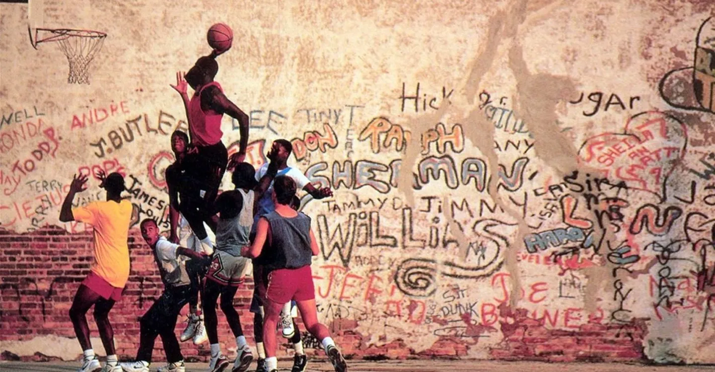 Scene from the 1989 “Michael Jordan’s Playground” poster, showing a high-flying dunk on an outdoor court with players gathered beneath and a graffiti-covered brick wall in the background.